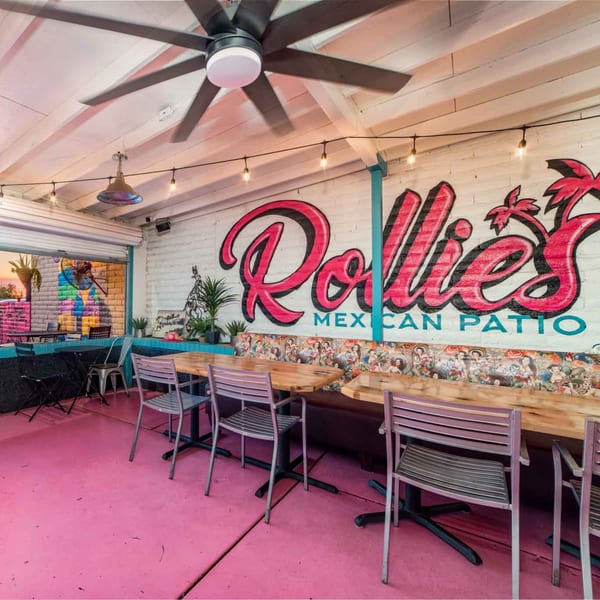 Rollie's Mexican Patio with pink floors, wooden tables, and a vibrant mural on a white brick wall.