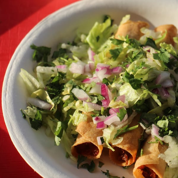 Crispy taquitos topped with shredded lettuce, chopped red onions, and cilantro on a white plate.