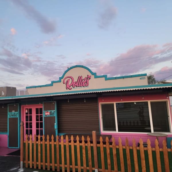 Exterior of Rollies Mexican Patio, a vibrant pink and teal building with a wooden fence under a cloudy sky.