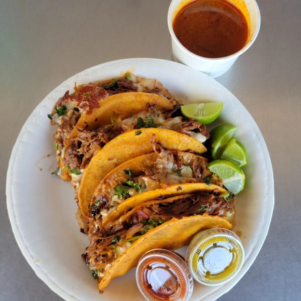 A plate of five birria tacos with melted cheese, cilantro, lime wedges, and a side of consommé.