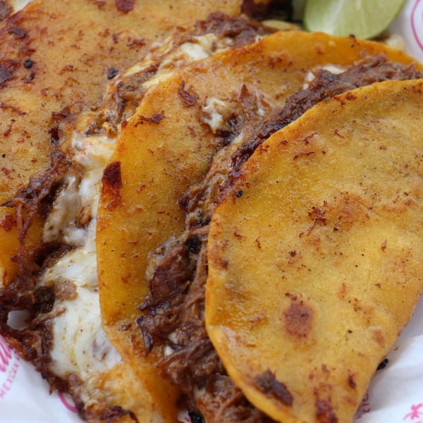 Close-up of three cheesy birria tacos with shredded meat, served with lime wedges.