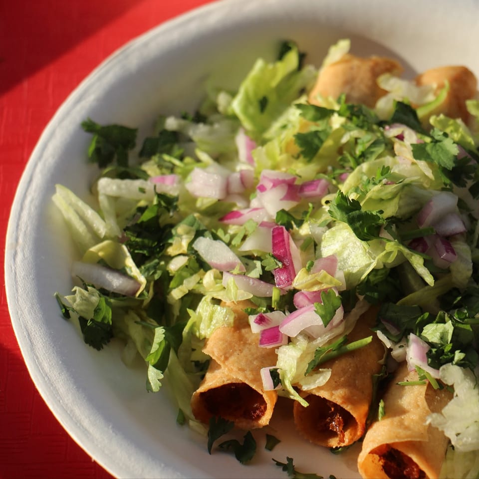 Crispy taquitos topped with shredded lettuce, chopped red onions, and cilantro on a white plate.