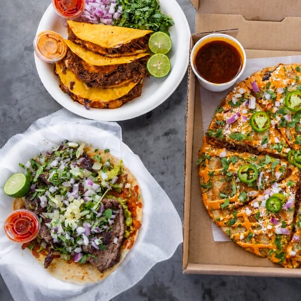 A spread of Mexican food, including birria tacos, a large carne asada taco, and a quesadilla pizza.